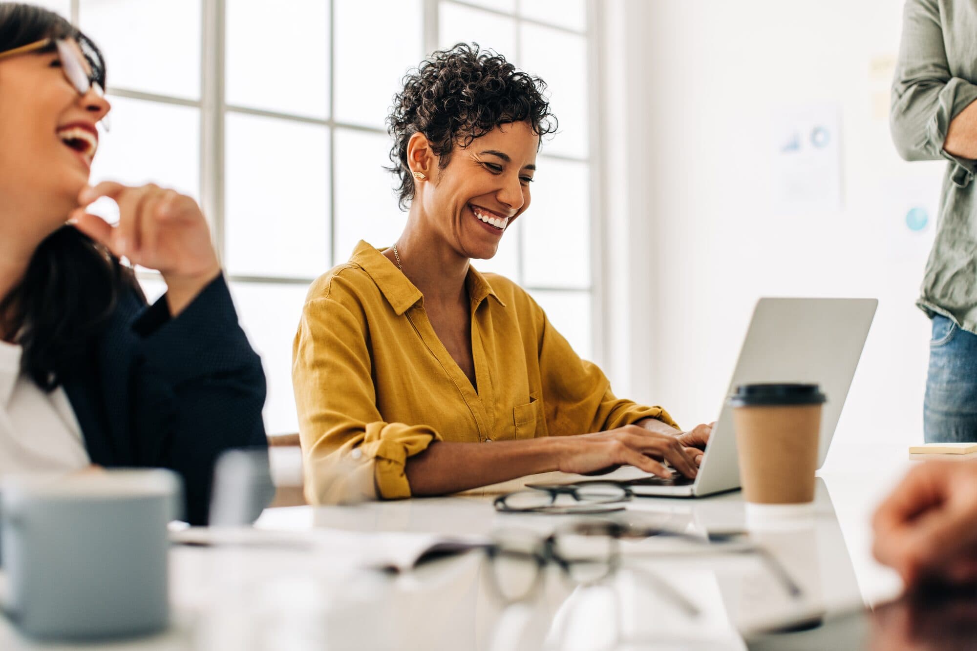 Colleagues laugh together at a meeting table with laptops and coffee