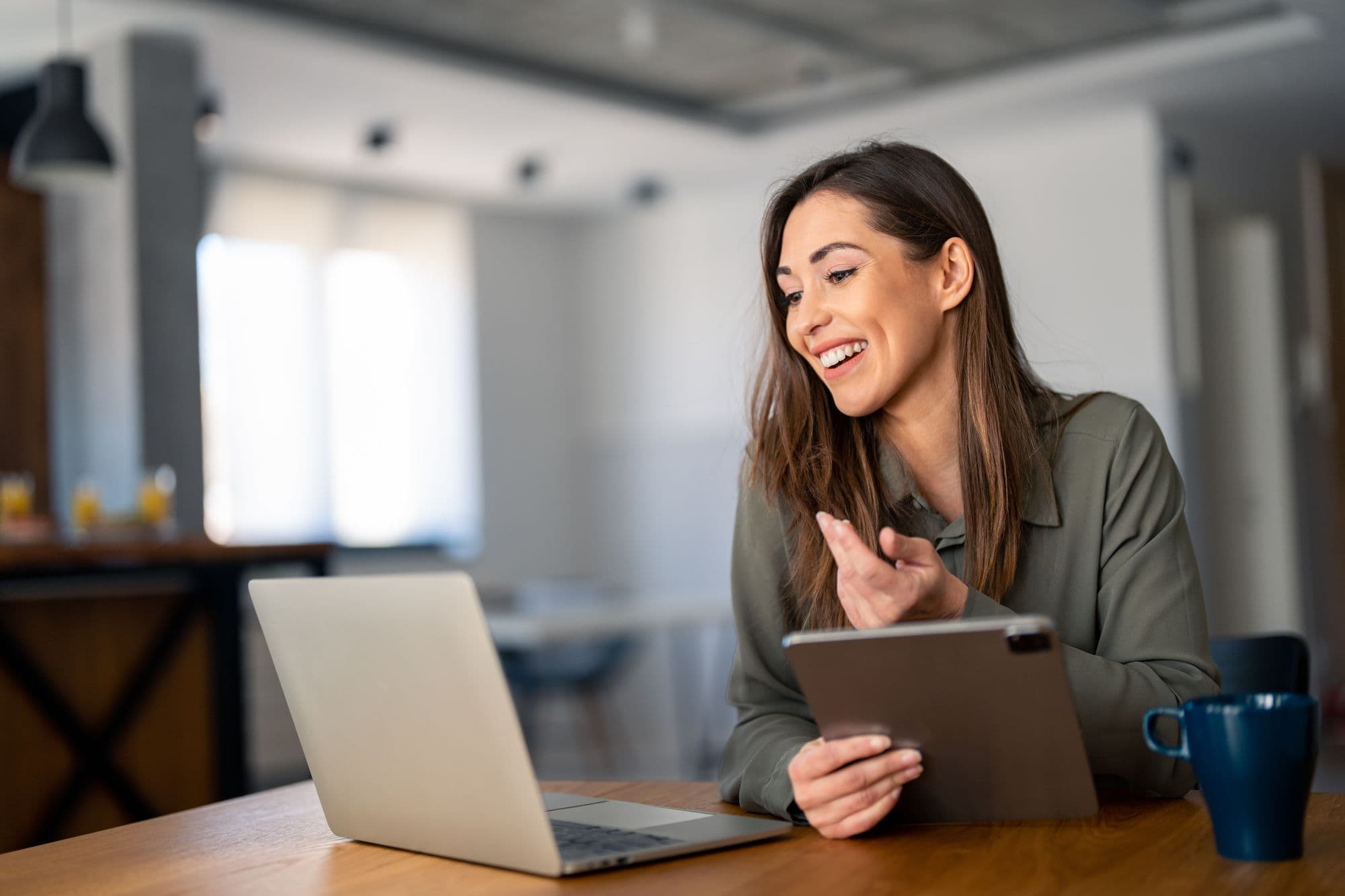 A woman laughs at her laptop, a tablet in hand