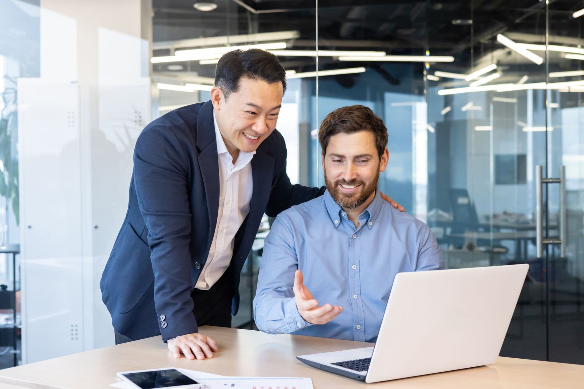 An experienced colleague leans over a coworker’s laptop as they talk and smile
