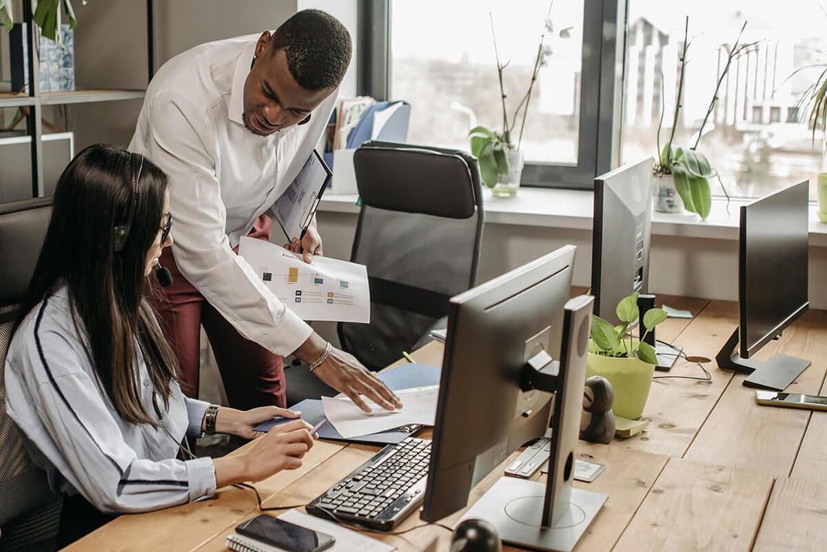man stands over woman at a communal desk in an office providing feedback on data and analysis of the mentoring program