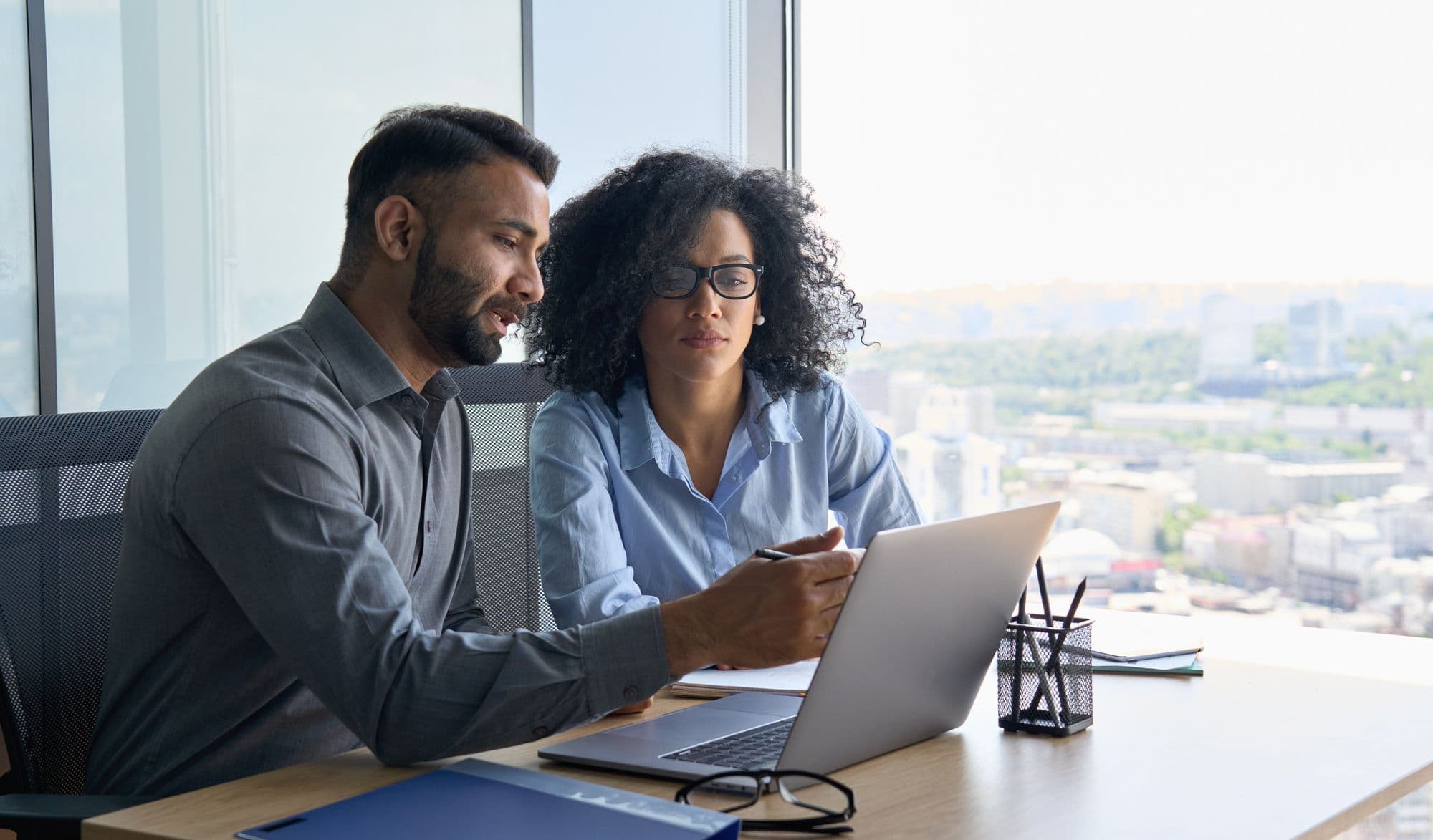 A man and a woman review a laptop by an office window with a city view