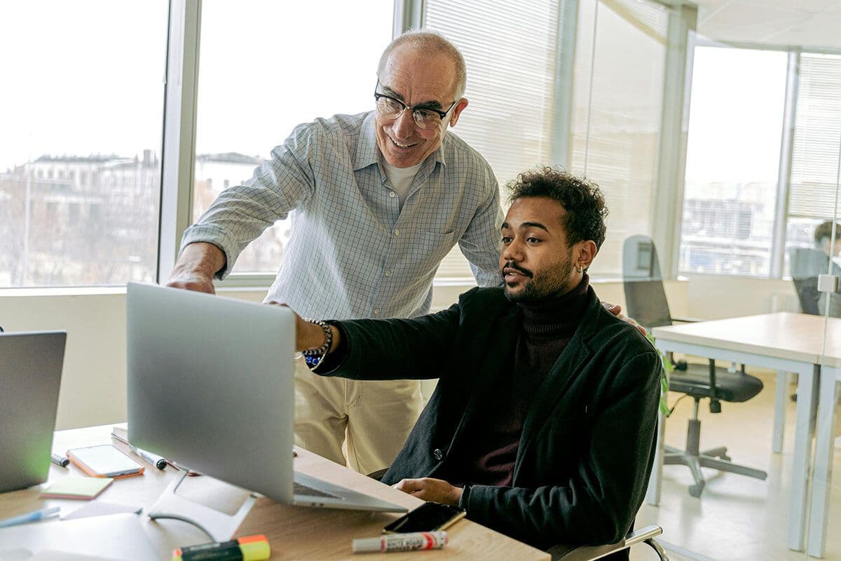 Two men in a modern office working at a desk on a laptop. Tee older man points to the screen, showing the younger colleague the 3 C's of mentorship.
