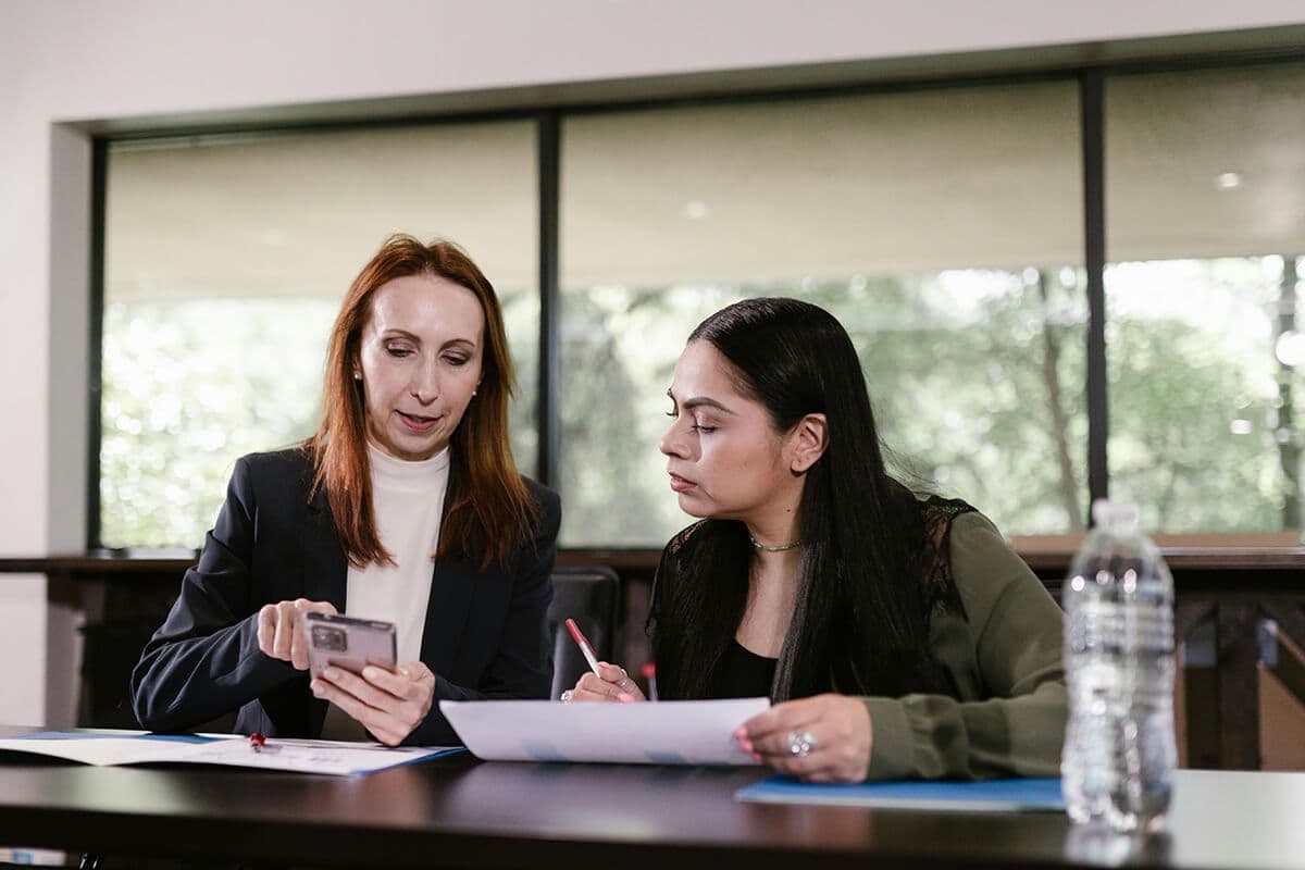 Two women sit at a desk reviewing papers and a mobile phone. The women are in a coaching session.