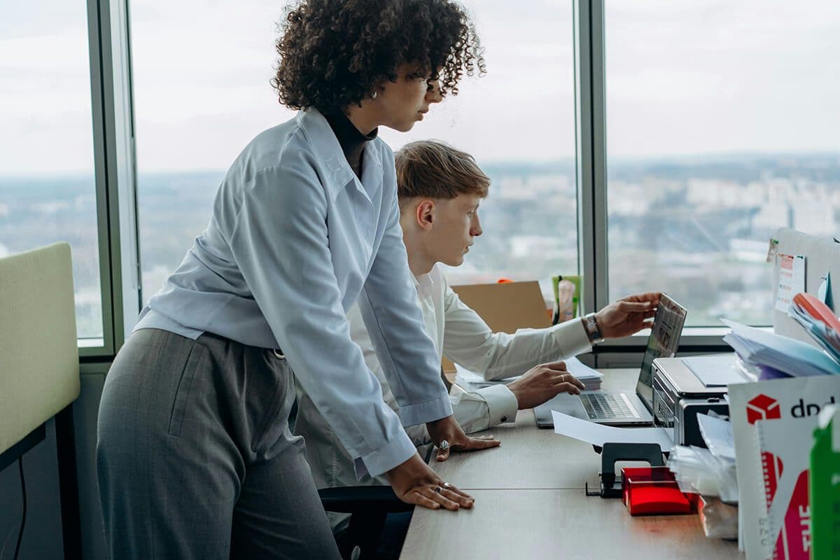 woman standing in office looking over the shoulder of male colleague staring at his laptop discussing the lack of collaborative culture at their organization.