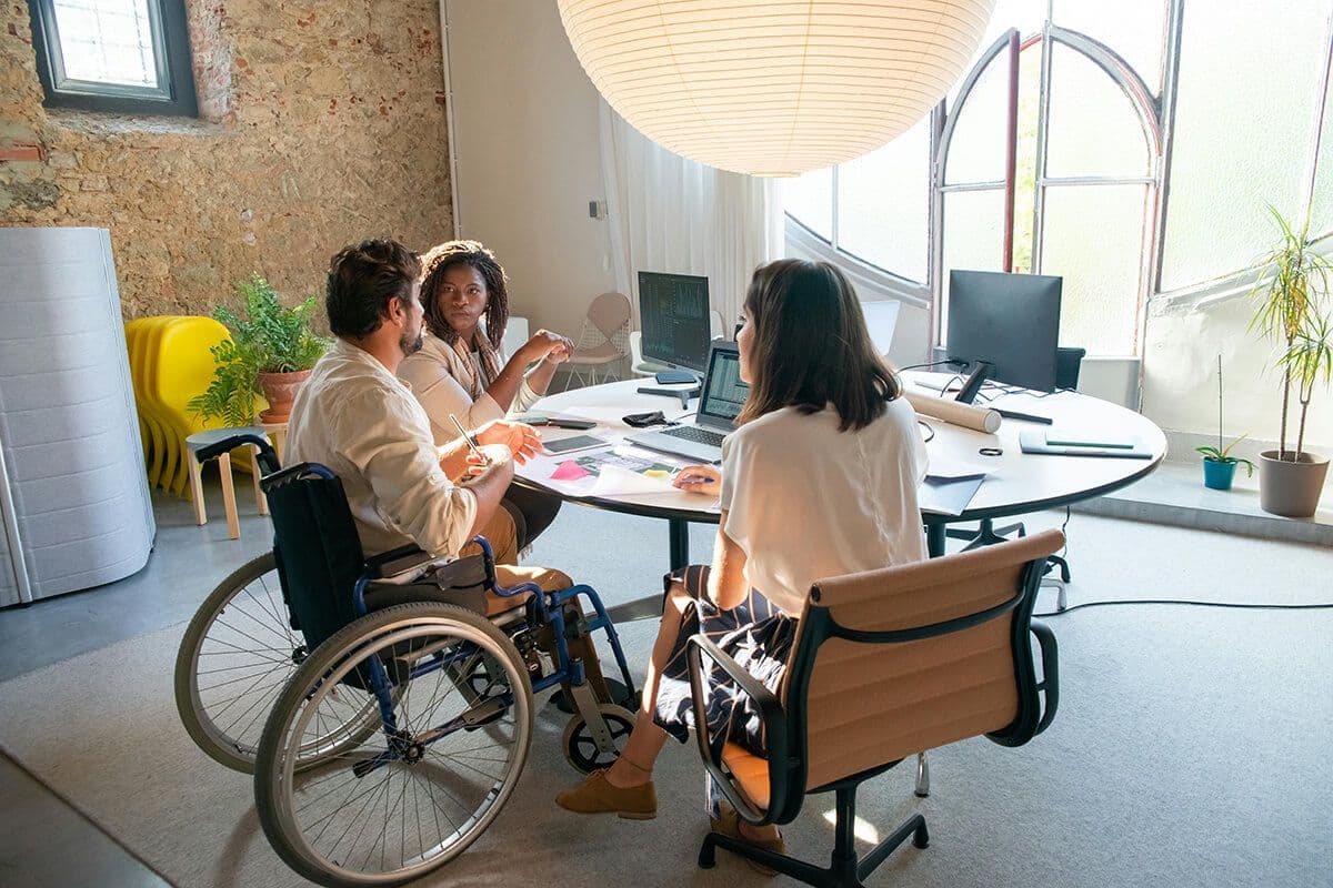 Two women employees sitting at a table with a male disabled colleagues, discussing performance and metrics at a table of computers, post-ti notes and paper.