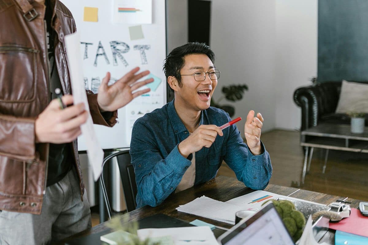 Asian man sits in modern office smiling while sitting next to a colleague giving a presentation on leadership styles.
