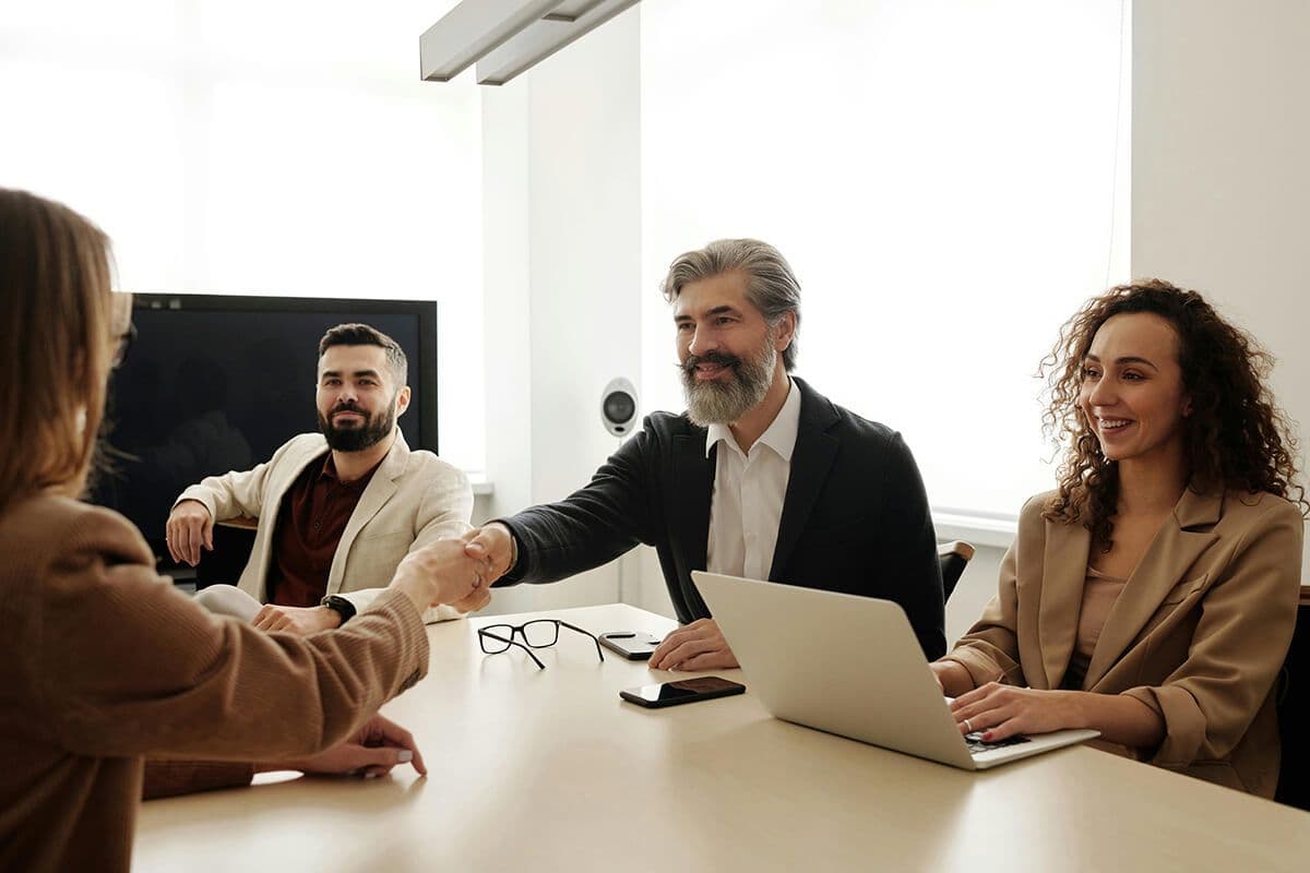People sitting in a conference room. Man with gray hair extending hand to woman across the table, while a woman sits on her laptop next to them. They discuss the benefits of varying leadership styles.